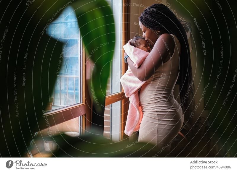 African American woman kissing newborn baby on hands child tender mother care love window home hug parent comfort embrace towel bonding apartment wrap
