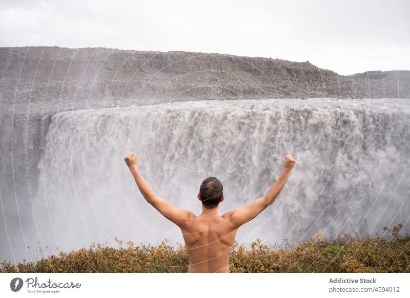 Shirtless man enjoying freedom near waterfall traveler success celebrate achieve adventure fist up viewpoint male admire activity shirtless goal rapid climb