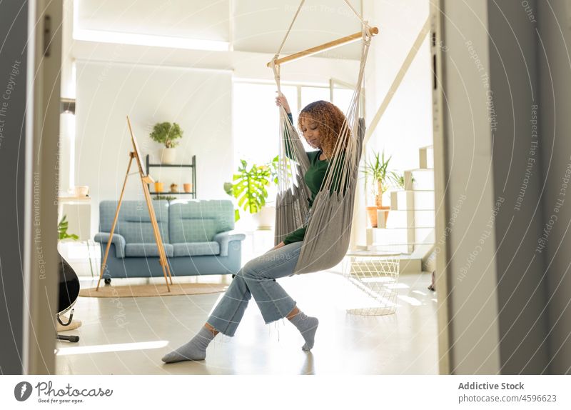 Young ethnic woman sitting on hammock in cozy living room comfort rest relax chill home enjoy casual free time recreation calm curly hair leisure peaceful