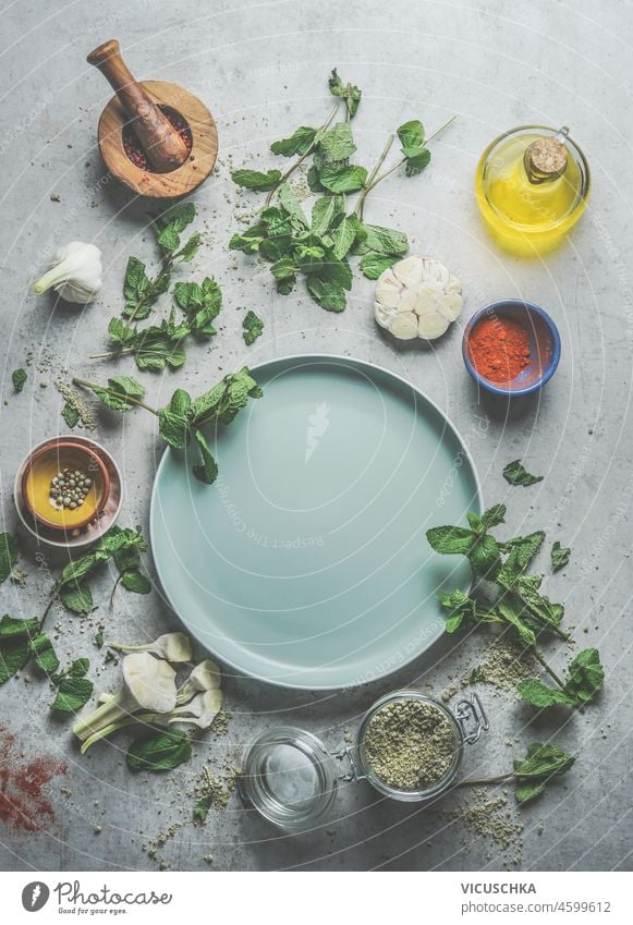 Various fresh seasoning and flavor ingredients around empty green plate with mortar and pestle on grey concrete kitchen table background. Flavorful cooking. Top view with copy space. Frame