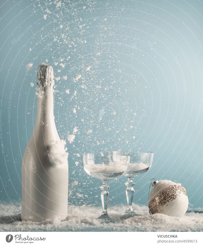 White champagne bottle and glasses with Christmas bauble on table with snowfall at blue wall background. New years concept with drink and falling snow. Front view.