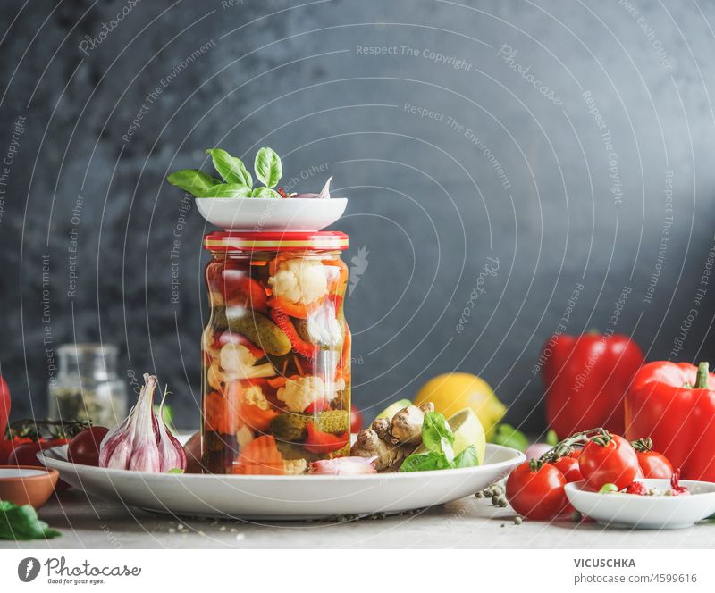 Homemade preserved vegetables in glass jar on white plate at table with kitchen utensils and ingredients, herbs and vegetables at black wall background. Healthy fermented food. Front view.