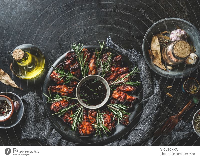 Marinated fried chicken wings with sauce and rosemary on dark plate at rustic black table with oil, spices and tablecloth. Top view with copy space. marinated