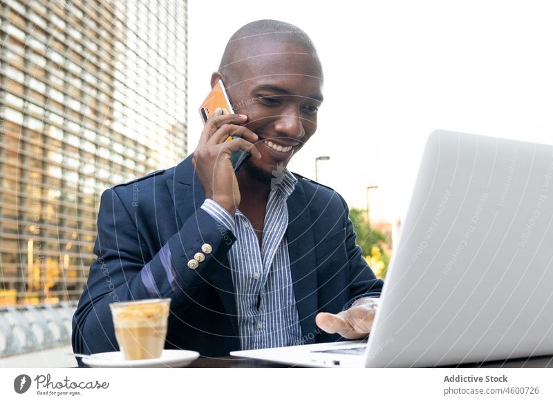 Gleeful black businessman talking on smartphone near laptop entrepreneur phone call conversation cafe terrace online street work male cup smile netbook job