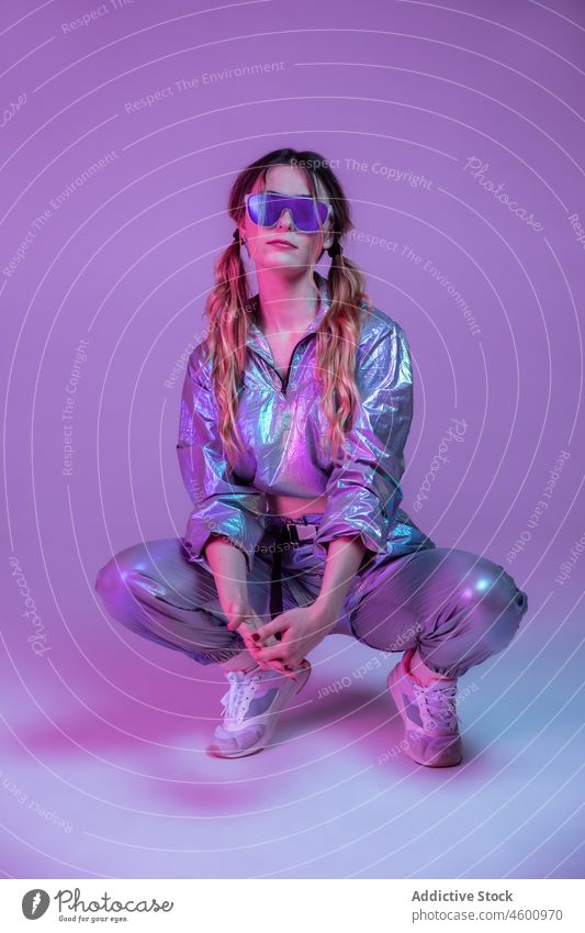 Trendy female in sunglasses crouched in neon studio woman model illuminate appearance trendy apparel personality 80s style futuristic young cloth individuality