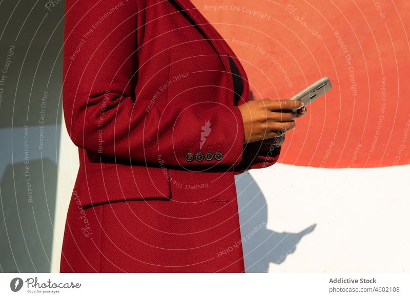 Anonymous African American woman in formal jacket with smartphone gadget cellphone using device street browsing red sunlight chat casual surfing social media