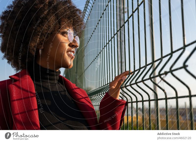 Black woman with Afro hairstyle looking away through metal grid afro fence street formal confident appearance outfit curly hair trendy individuality eyeglasses