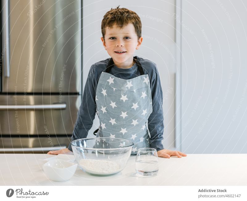 Smiling boy with bowl of flour in kitchen cook culinary kid ingredient home domestic child prepare table apron cheerful food smile positive pastry glass water