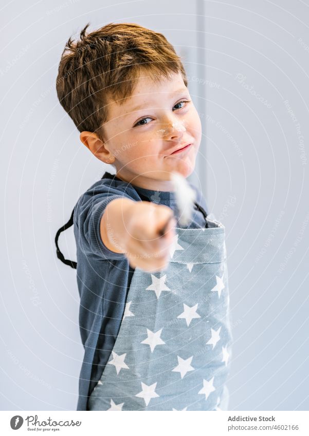 Cute boy with spoon cooking in kitchen kid helper domestic child kitchenware culinary at home apron dirty utensil funny smile prepare messy brown hair flour