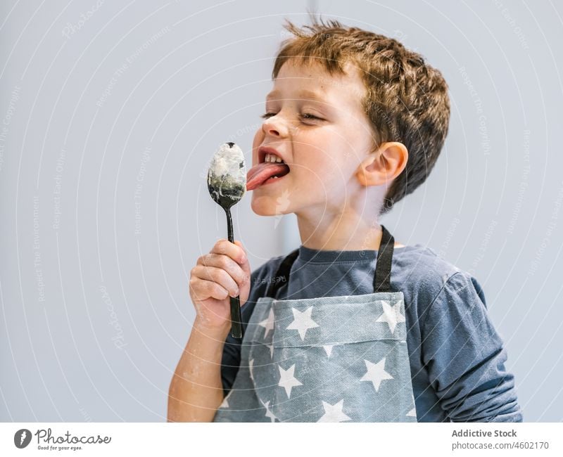 Boy in apron licking spoon with dough boy cook kid culinary domestic child kitchenware helper at home dirty utensil funny prepare messy eat brown hair flour