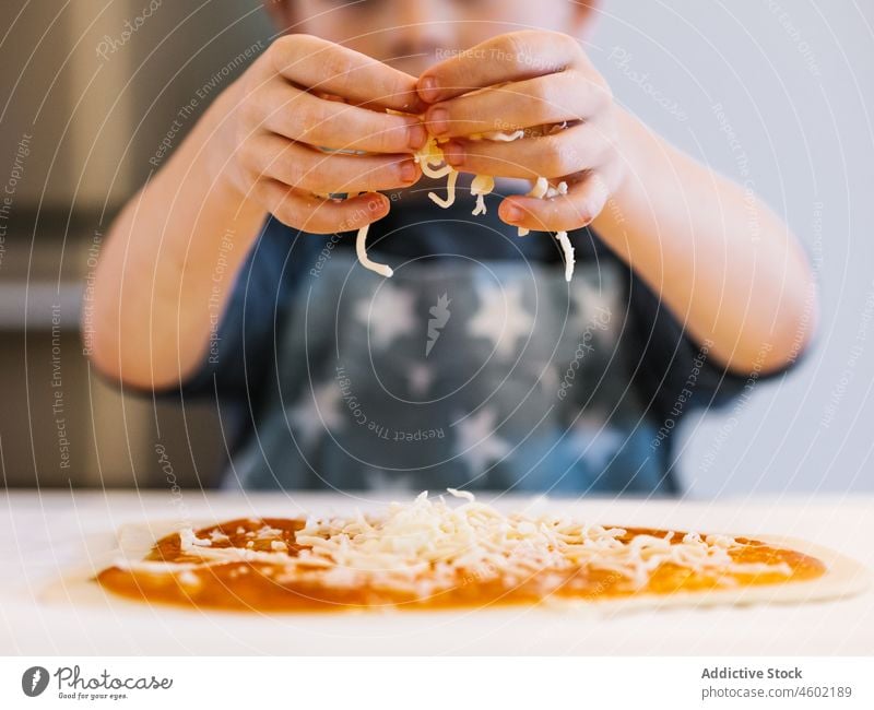 Anonymous boy putting cheese on pizza cook kid culinary kitchen dough child helper kitchenware domestic at home apron recipe food cuisine add prepare process