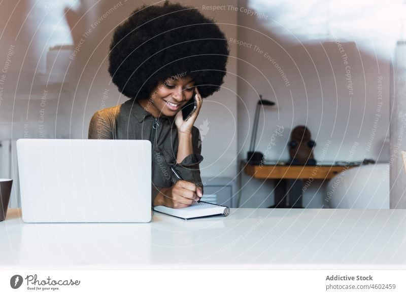 Happy black woman writing in notebook near laptop work freelance workplace phone call take note speak online agenda smartphone schedule mobile phone modern