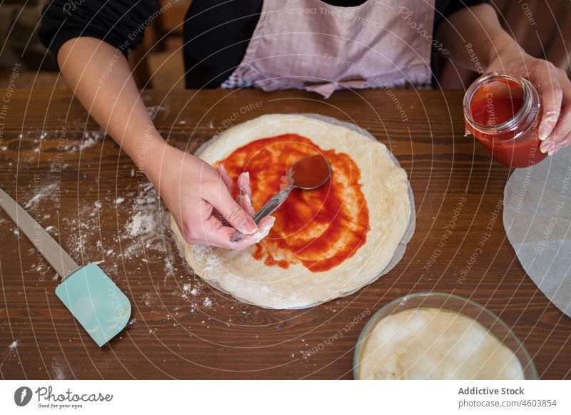 Anonymous woman adding sauce on dough cook pizza tomato kitchen culinary homemade raw cuisine prepare chef table apron process female light lady housewife flour
