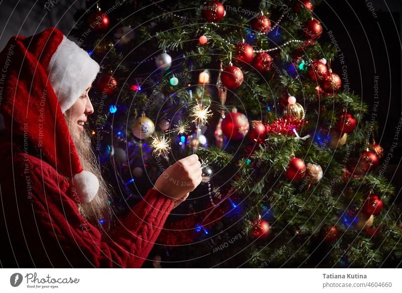 woman in a Santa hat with lighted sparklers on the background of a New Year or Christmas tree decorated with red ball. celebrate new year christmas party happy