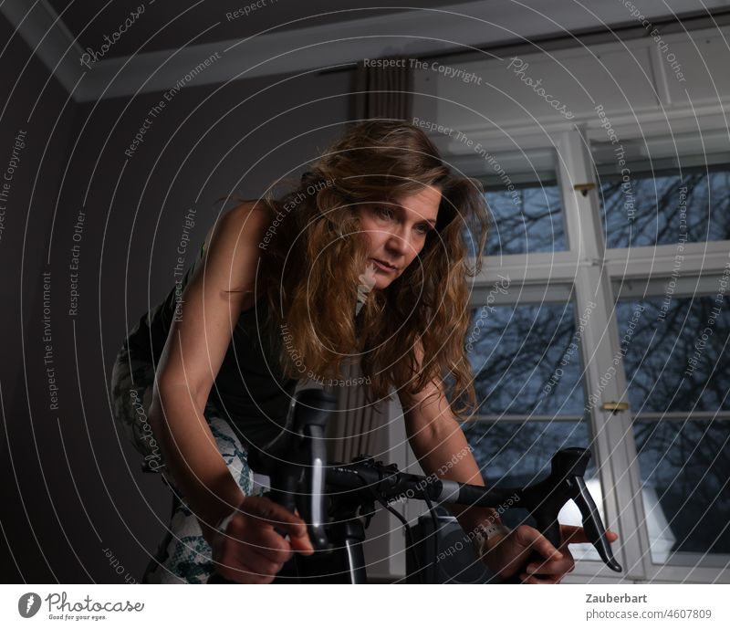 Sporty beautiful woman training on indoor racing bike, behind window with evening light Woman Athletic pretty exercise Racing cycle good resolutions