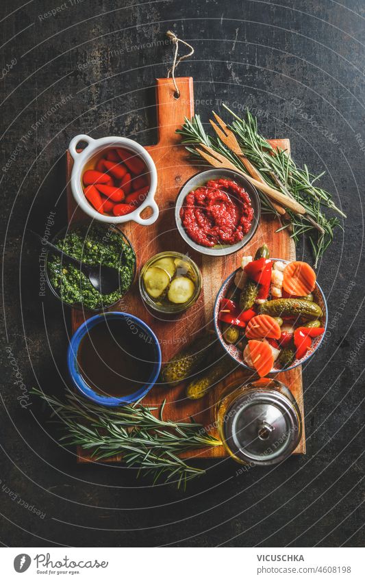 Bowls with various pickled vegetables snack on wooden cutting board with fresh herbs, pesto and homemade ketchup on dark rustic background. Top view bowls