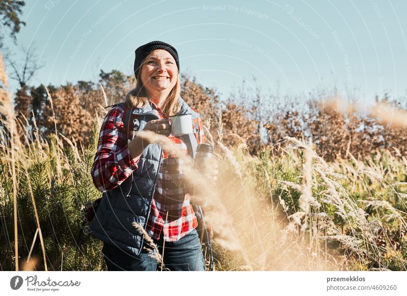 Smiling woman relaxing and enjoying the coffee during summer trip. Woman standing on trail and looking away holding cup of coffee. Woman with backpack hiking through tall grass along path in mountains
