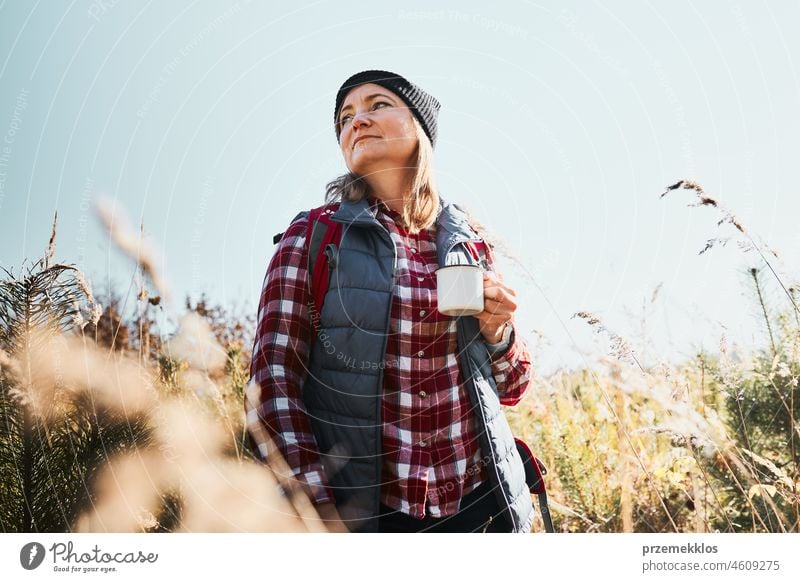 Woman taking break and relaxing with cup of coffee during summer trip. Woman standing on trail and looking away. Woman with backpack hiking through tall grass along path in mountains