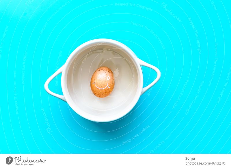An egg with smiling face in a cooking pot on a blue background. Top view. Hen's egg Brown Smiling Breakfast saucepan Protein Humor Organic produce Eggshell