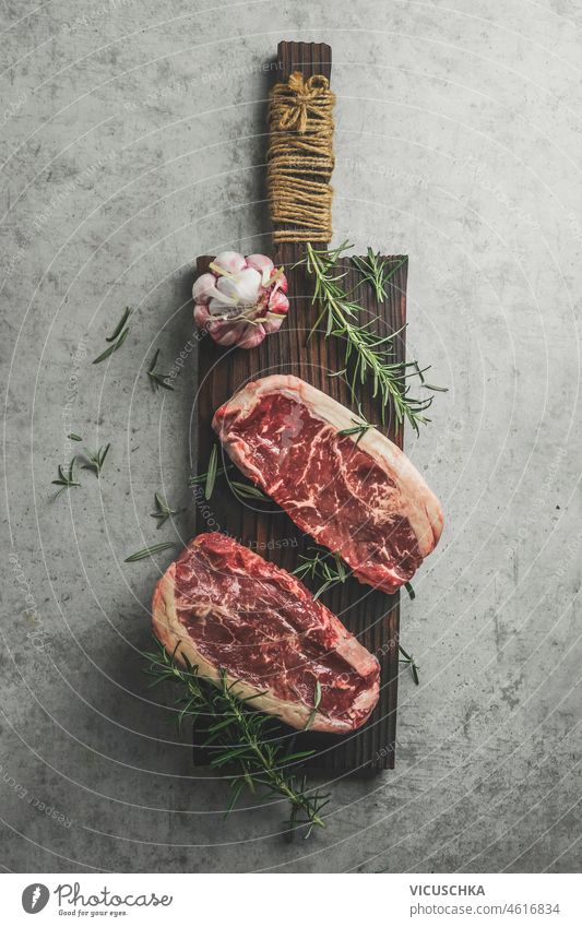Two raw marbled rib-eye steak on cutting board with rosemary, garlic and green pepper rib eye two marbled steak concrete kitchen table barbecue preparation