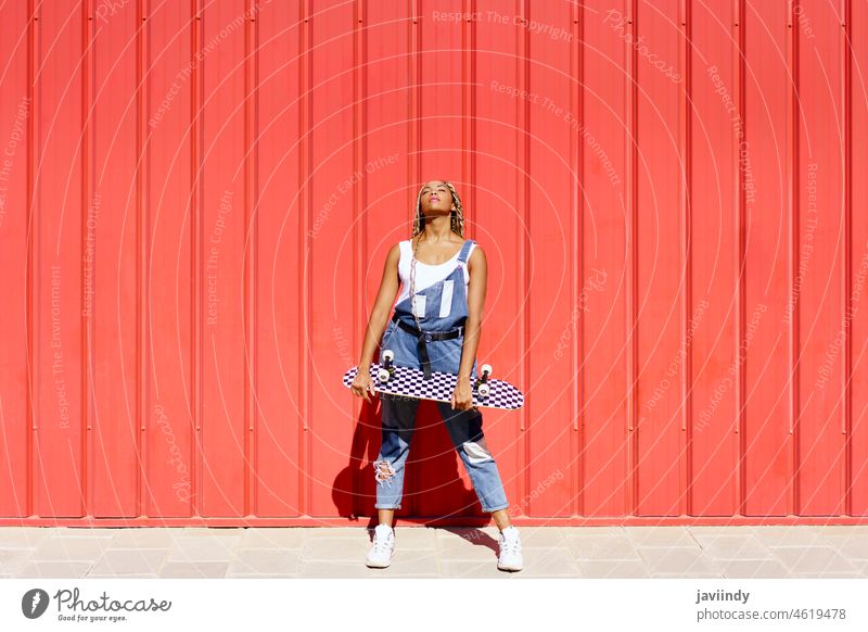 Black girl dressed casual, wtih a skateboard on red urban wall background. black braids afrobraid dreadlocks woman african hairstyle young female person outdoor