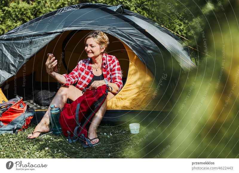 Female having video call with friends using smartphone while sitting in tent at camping. Woman relaxing in tent during summer vacation trip adventure campsite