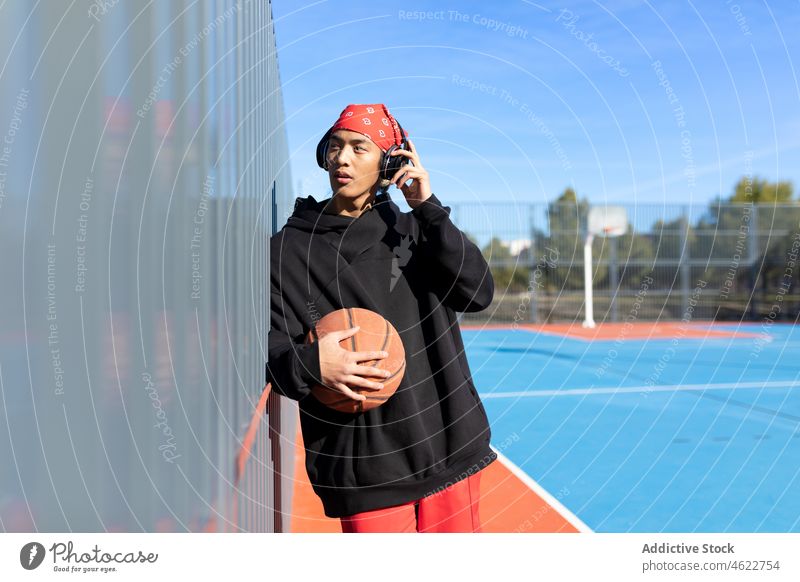 Asian man with basketball ball listening to music on playground headphones song portrait sport game hobby training asian fence sports ground male activity