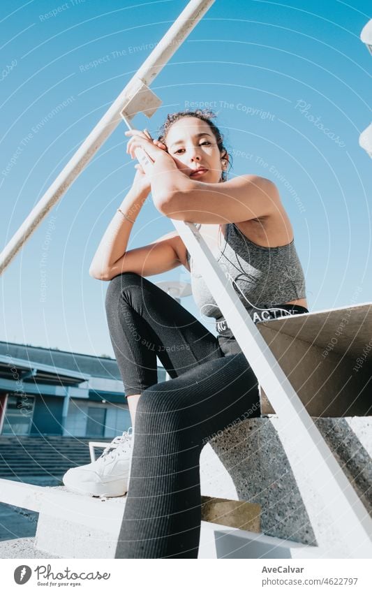 Young arab african woman on training sport wear leggins and top sitting on stairs while listening to music resting after workout. Serious looking to camera,beauty young athlete urban city background