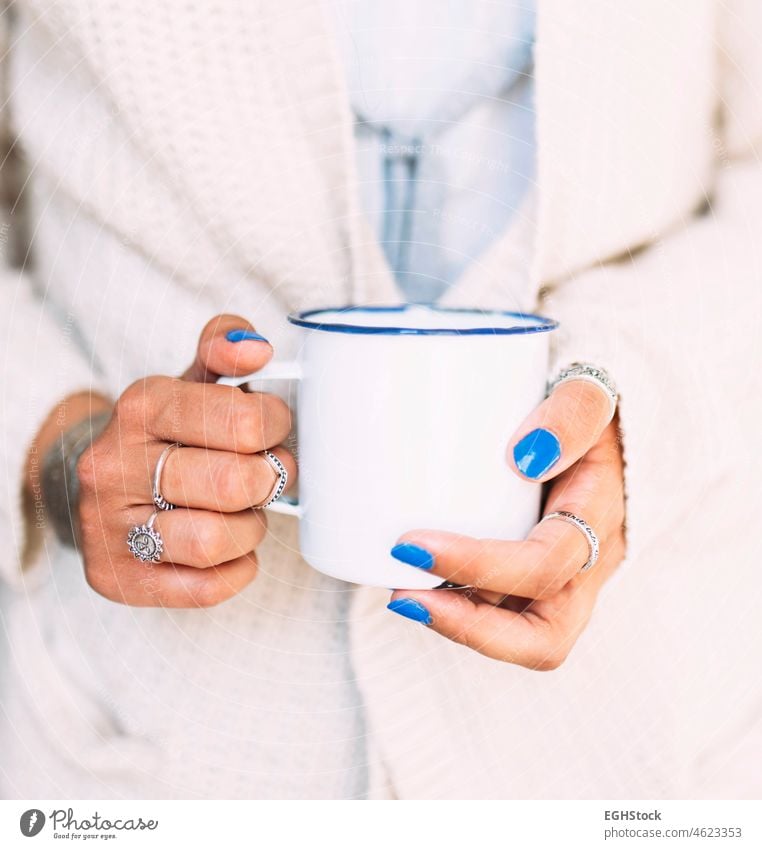 Close view of the hands of a young woman with blue painted nails and finger rings holding a white enameled mug with coffee or tea cup finguers winter jumper