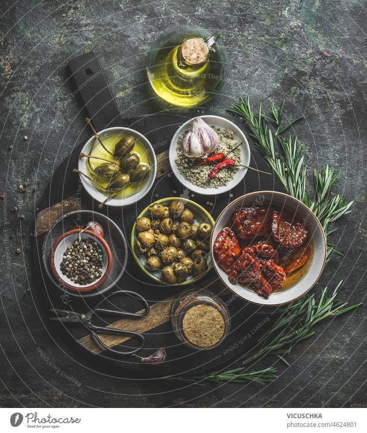 Various tasty Mediterranean food appetizer in bowls : olives, capers, dried tomatoes , fresh rosemary herbs and olive oil served on rustic cutting board various