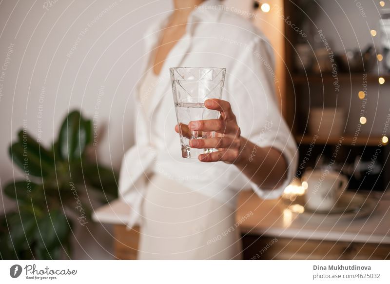 Unrecognizable woman in white shirt holding a glass of water, standing in the kitchen with evening light. Healthy lifestyle - drinking enough water every day