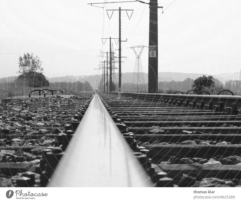 Railroad track with power poles in black white Railway rail Electricity pylon Black/White stones Transport Environment Climate country