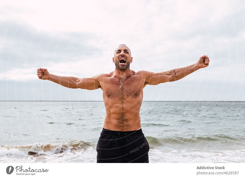 Shirtless man with spread arms on beach naked torso shirtless sea training water shore healthy lifestyle wellbeing sporty muscular male scream workout seashore