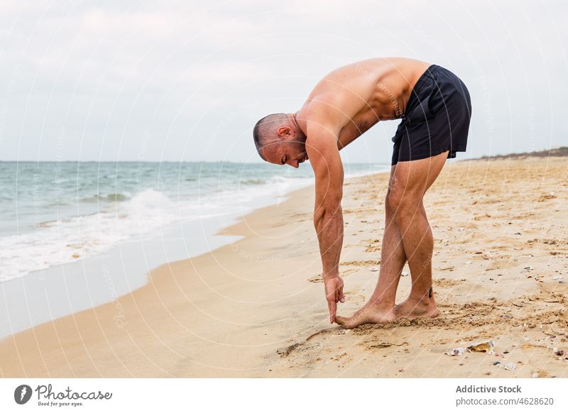 Shirtless man stretching and bending forward on beach shirtless sea training water shore healthy lifestyle exercise practice sporty wellbeing muscular male