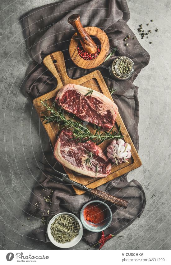 Raw marbled steaks with seasoning in bowls , rosemary and kitchen utensils raw wooden cutting board tablecloth grey background cooking fresh meat top view