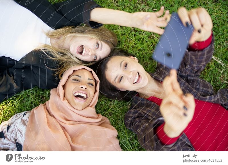 Cheerful diverse girlfriends laughing and taking selfie lying on lawn in park women smartphone together best friend spend time friendship mobile grass excited