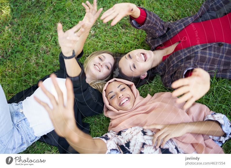 Cheerful young diverse female friends lying on grass and smiling at camera women smile lawn park together wave hand friendship optimist happy greeting