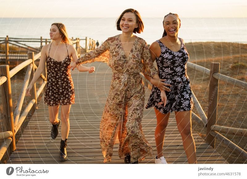 Smiling multiethnic girlfriends walking on wooden boardwalk on sea shore women joy cheerful sunset style coast happy female holding hands together stroll enjoy