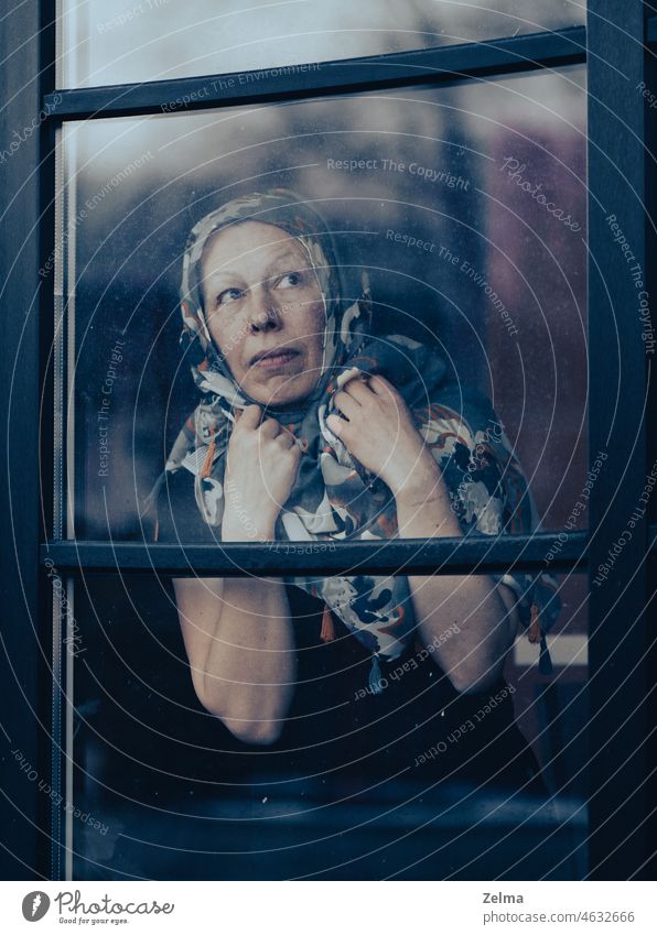 Sad mature woman with scraft on head standing behind dirty window glass middle-aged elderly look through scarf face expression hands indoor old senior disease