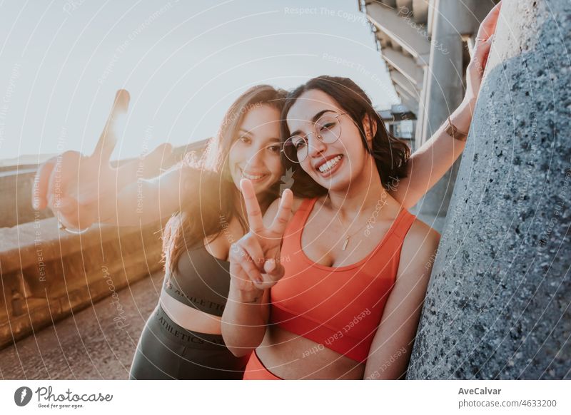 Two Young cheerful sportswoman training outdoors doing exercise smiling with happy at the camera doing victory sign with fingers during a colorful sunset. Working out together concept. Healthy life