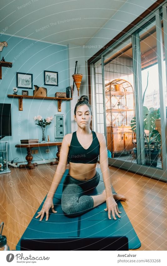 Attractive fitness young woman sitting in Half Pigeon Pose doing yoga with online class at home. Entertainment and education on the Internet. Healthy lifestyle concept during free time.