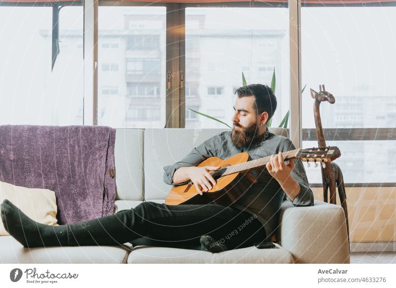 Young handsome bearded Man playing acoustic spanish guitar sitting on the sofa in the living room. Learning to play a new instrument. New habit concept, skills and hobby. Modern home trendy clothes