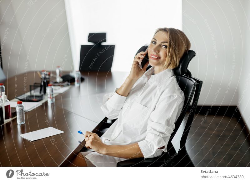 A businesswoman is talking on the phone sitting at a desk in the office mobile looking smiling window modern person professional indoors owner smartphone