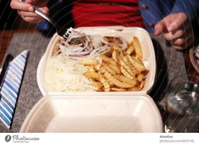 Woman with red sweater and blue jacket sorts onion strips from a styrofoam menu box from a gyro plate with fries and coleslaw - Dinner from the delivery service - The small Kollossos- plate