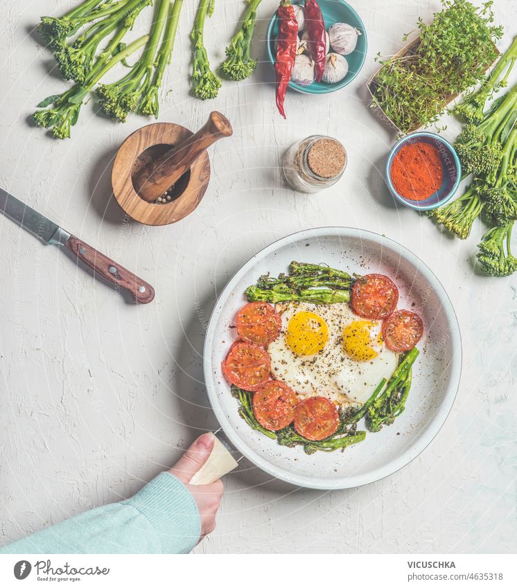 Women hand holding white pan with homemade breakfast: fried eggs, broccoli, tomatoes and spices on pale grey kitchen table with mortar, pestle and ingredients. Low carb food. Top view.