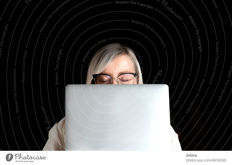 tired mature woman's face with closed eyes and glasses hidden behind a computer laptop isolated on a black background eyeglasses home work female using