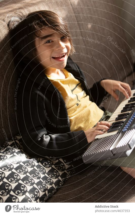 Cheerful ethnic boy playing on keyboard kid synthesizer instrument childhood musical song melody entertain living room home appearance domestic apartment light