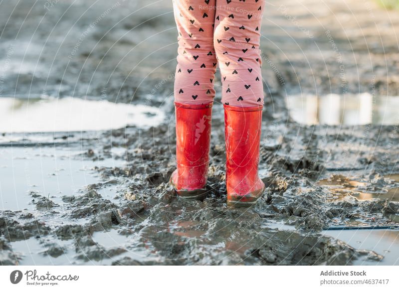 Anonymous kid standing on dirty ground pathway gumboots mud puddle wet water street childhood rubber sidewalk color walkway bright footwear surface road leg red
