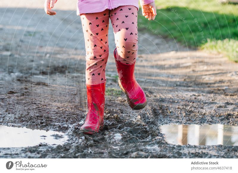 Anonymous kid standing on dirty ground pathway gumboots mud puddle wet water street childhood rubber sidewalk color walkway bright footwear surface road leg red