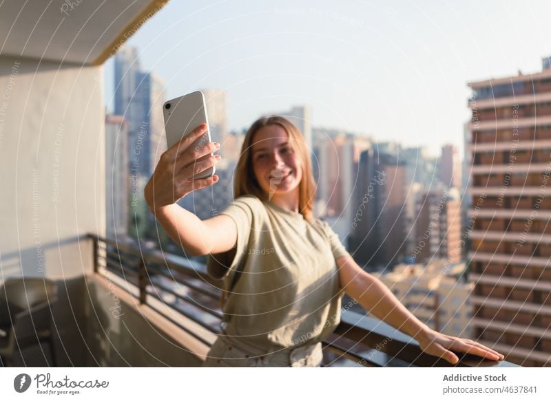 Happy woman taking selfie on balcony with city view smartphone smile terrace building positive traveler holiday mobile architecture female young long hair